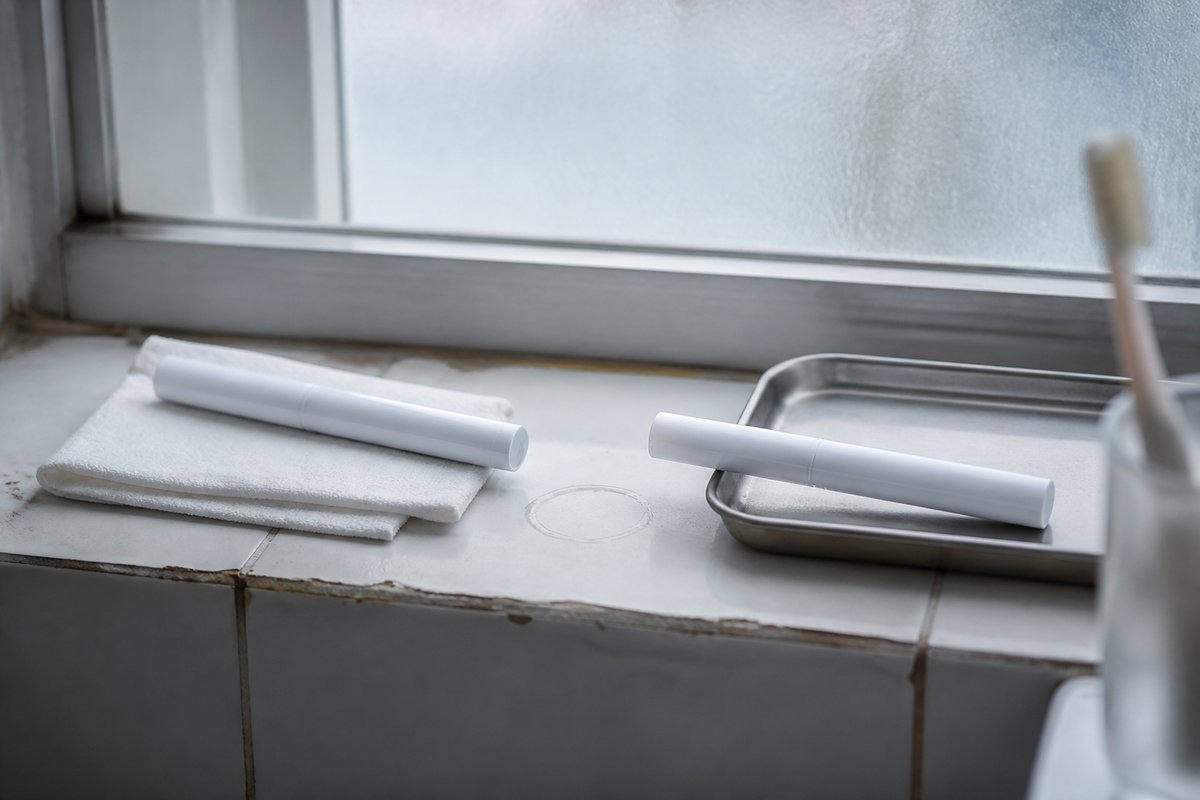 A still life of two objects standing apart on a pale bathroom shelf: a slim reusable pen-shaped injector lying on a fold